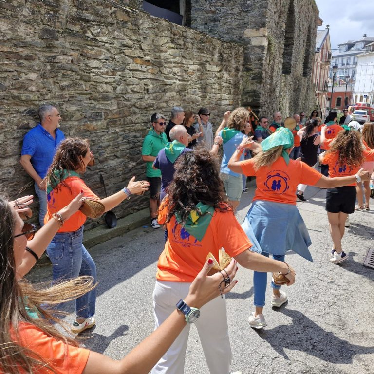 Varias personas practican baile tradicionalen la calle con la Muralla de Navia al fondo.